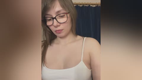 Video of an Asian woman with straight brown hair, wearing glasses, a white tank top, and looking down in a dimly-lit room with dark curtains.