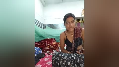 A video of a young South Asian woman with medium skin tone and dark hair, wearing a black blouse and floral-patterned skirt, sitting on a bed with green and red covers in a modest bedroom.