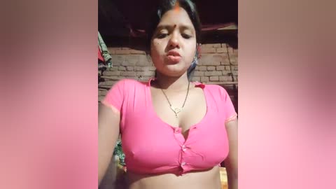 Media: Video of a young woman with medium brown skin, dark hair, and medium breasts, wearing a pink crop top, in a dimly lit room with brick walls.