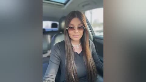 Video of a Latina woman with long brown hair, wearing sunglasses, black top, and necklace, seated in the backseat of a car, looking relaxed.