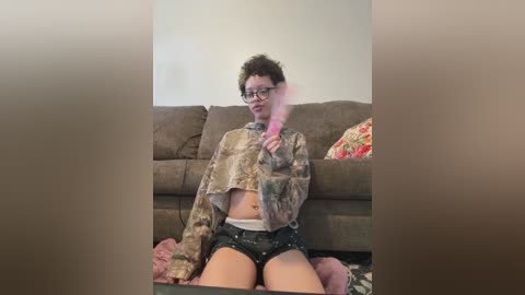 Video of a young, slender woman with short curly hair, wearing glasses, a camo jacket, and shorts, sitting on a couch, blowing pink smoke from a vape.