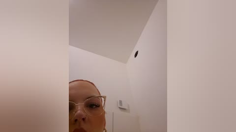 Video of a young woman with fair skin and brown hair, wearing clear glasses, standing in a narrow, white-walled corridor with a small, dark hole above her head.