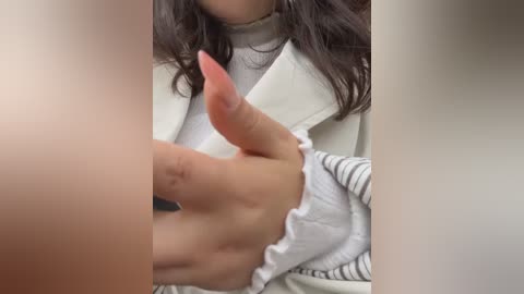 A close-up video of a person with fair skin, wearing a white sweater with a frilled collar, holding their erect penis with a pink nail polish. The background is blurred and indistinct.