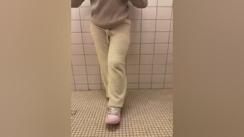 Video of a person in a beige sweater and light cream pajama pants, standing on a beige tiled floor in front of white-tiled walls. The person's feet are in white sneakers.
