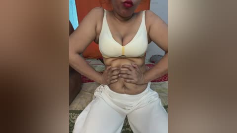 Video of a woman with medium skin tone, wearing a yellow bra and white pants, sitting on a patterned bed, hands on her stomach, red lips, blurred background.