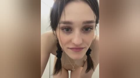 A close-up video of a young Caucasian woman with pale skin, brown pigtails, and a slight smile. She is topless, revealing small breasts. The background is blurred, suggesting a bathroom setting.