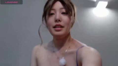 A video of a young Asian woman with fair skin, wearing a light purple spaghetti-strap top, standing indoors with a slightly blurry background. She has shoulder-length brown hair and is smiling slightly.