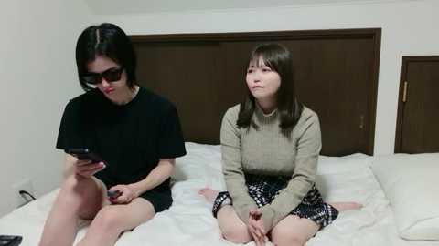 A video shows two young women, one with short black hair and sunglasses, the other with shoulder-length brown hair, sitting on a white bed in a simple room.