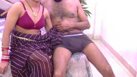 Video of a shirtless South Asian man wearing black boxers, seated next to a woman in a maroon bra and striped skirt, both indoors on a tiled floor.