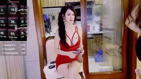 A video of an Asian woman with long black hair in a red, revealing, low-cut dress sitting on a white chair in a room with a TV displaying a weather forecast.