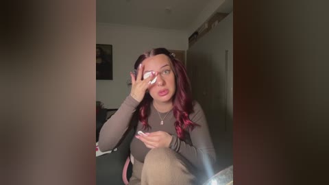 A video of a young woman with long, wavy, red hair, wearing a gray top, sitting on a black chair, covering her face with her hand, in a dimly lit room with a painting on the wall.