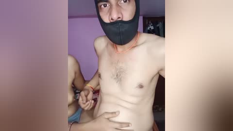 A video shows a young, light-skinned man with a black mask and orange necklace, his chest hair visible, holding his groin. Two hands, possibly female, touch his body. Background includes a purple wall and indistinct furniture.