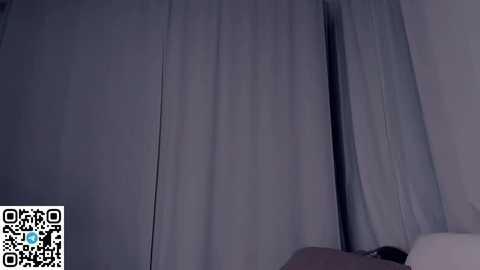 A dimly lit video shows a bedroom with a large, closed window partially covered by sheer, light gray curtains. The image includes a QR code in the bottom left corner.