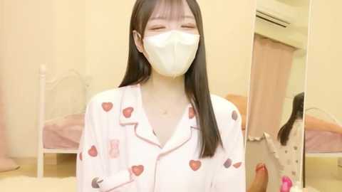 Video of an Asian woman with long black hair, wearing a white mask and a pajama shirt with strawberries, standing in a bright, minimalist bedroom with a bed and a mirror.