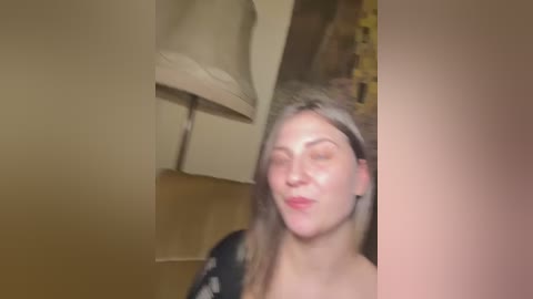 A blurry video of a woman with long, light brown hair, wearing a black robe, smiling with eyes closed, in a dimly lit room with beige walls and a lampshade.