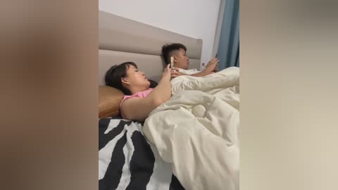 Media: Video of an Asian couple in bed, man with short dark hair, woman with straight black hair, both wearing white pajamas, zebra-patterned bedspread, beige headboard, white walls, daytime lighting.