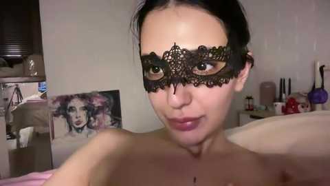 Video of a topless woman with fair skin and dark hair, wearing a black lace masquerade mask, in a softly lit bedroom with art on the wall and makeup on a table.