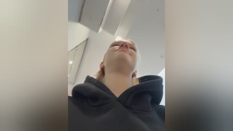 Video of a young woman with fair skin and blonde hair, wearing a black hoodie, looking upward in a white-walled room with a mirrored ceiling.