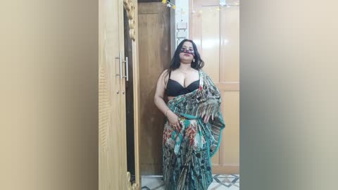 Video of a curvy, dark-skinned woman in a colorful sari, black bra, and sunglasses, standing in a hallway with wooden doors and beige walls.