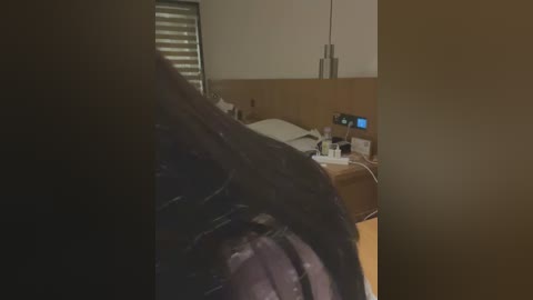 A video of a dimly lit hotel room featuring a partially visible person in black clothing, a wooden headboard, a bedside table with a lamp, and a digital clock. The room's ambiance is warm and muted.