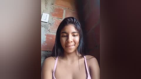 Media: A video of a young woman with medium brown skin, straight black hair, and a confident smile. She wears a light pink spaghetti-strap top, standing in front of a rustic brick wall with a white switch plate visible.
