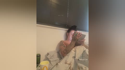 Media: A video of a woman lying in a bed, wearing a brown sweater, reading a tablet. The room has a white wall, a window with a closed blind, and a green can on a nightstand.