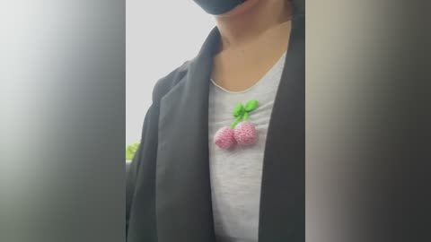 A close-up video of a person wearing a black mask, gray shirt with a pink strawberry patch, and a black blazer. Background is blurred, with hints of green foliage.
