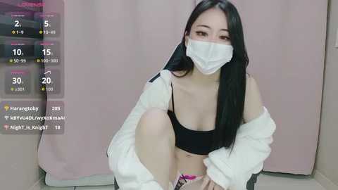 Video of a young Asian woman with long black hair, wearing a white mask, black bra, and white shirt, sitting on a bed, with a digital screen showing social media engagement metrics.
