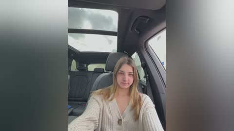 Media: Video of a young Caucasian woman with long blonde hair, wearing a white cardigan, seated in the driver's seat of a car. The car's interior is modern with black leather seats and a cloudy sky visible through the windshield.