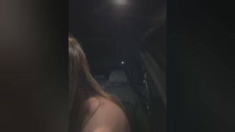 A dimly lit video of a woman with long brown hair seen from behind, sitting in a car. The scene is dark, with only a faint light source visible above.
