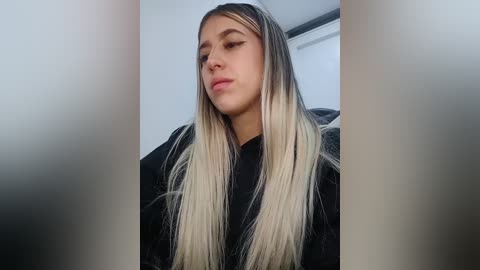 Video of a young woman with long, straight, platinum blonde hair, fair skin, and a neutral expression, wearing a black top. The background is a blurred, modern indoor setting with white walls.