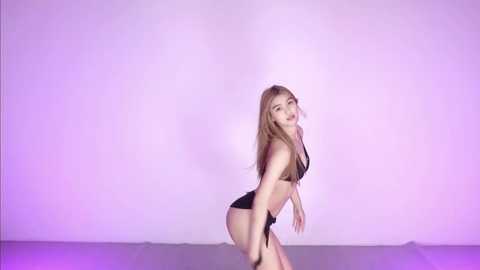 Video of a slender, fair-skinned woman with long, straight blonde hair, wearing a black bikini, posing in a studio against a gradient purple and pink backdrop.