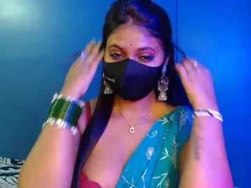 Video of a woman with medium skin tone, wearing a black face mask, green sari, and silver bracelets, adjusting her hair against a blue wall.