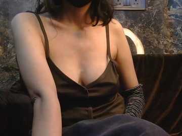 Video of a woman with medium skin tone, wearing a black camisole and black striped gloves, seated in a dimly lit room with a rustic, wooden backdrop.