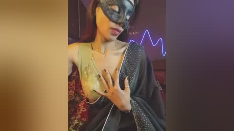 A video of a woman in a black saree with a gold blouse, wearing a black mask with a golden eye, holding her breast. The background features a dimly lit room with a neon heart sign.