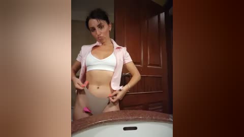 A video of a slim, fair-skinned woman with short dark hair, wearing a white sports bra and pink shirt, standing in a bathroom, adjusting her white panties.