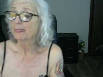 A video of an older woman with shoulder-length, wavy white hair, wearing glasses and a black tank top, with a colorful tattoo on her left arm. She sits in a black office chair against a plain, gray wall.
