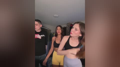 Media: Video of three young adults in casual attire: a man in a black shirt, a woman in a black sports bra and yellow pants, and a woman in a black sports bra and grey pants. Background shows a dimly lit room with curtains.