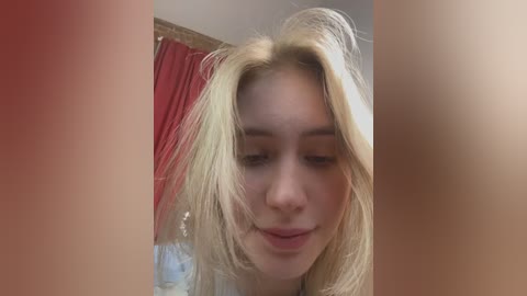 Video of a young woman with long, blonde hair, fair skin, and a slight smile, standing in a room with red curtains and a wooden frame.