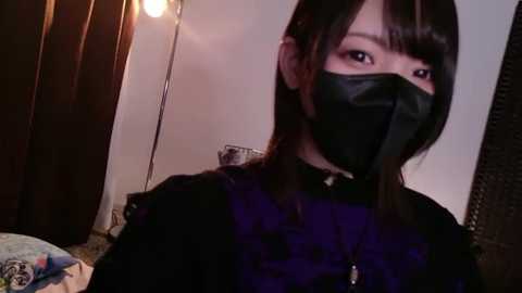 Video of an Asian woman with straight black hair and a black face mask, wearing a black dress, standing indoors with a dimly lit room in the background.