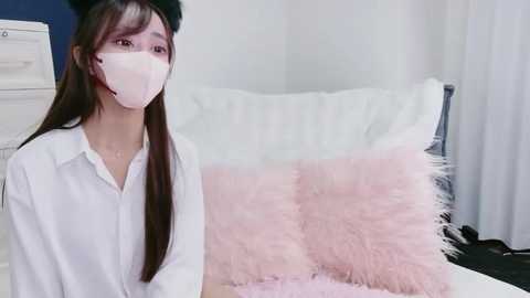 Video of a young Asian woman with long dark hair, wearing a white button-up shirt, a black beret, and a white face mask, sitting on a bed with a fluffy pink pillow.