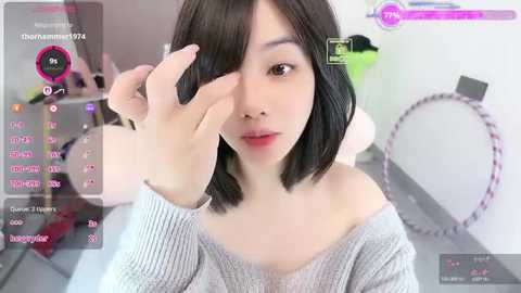 A young Asian woman with short black hair and fair skin, wearing a grey off-shoulder sweater, applies makeup on her right eye in a bright, modern room.