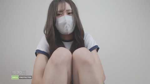 Video of a young Asian woman with long, straight brown hair, wearing a white short-sleeved shirt and a white face mask, crouched with knees up, against a plain white background.