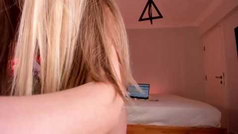 Media: Video of a blonde woman with straight hair in a dimly lit bedroom, sitting at a bed with a laptop open on a white sheet.