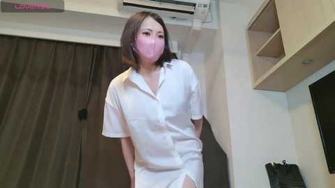 Video of an East Asian woman with medium-length black hair and a white surgical mask, wearing a white button-down shirt, standing in a modern, minimalistic room with beige walls, a TV, and dark curtains.