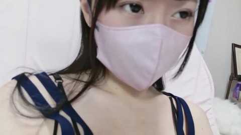 Media: Video of a young Asian woman with long black hair, fair skin, and light pink face mask, wearing a blue and white striped top, indoors with a white wall background.