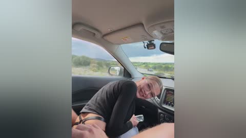 Video of a man in a car, smiling while adjusting his pants, wearing glasses and a black shirt. Outside, there's a blurred landscape.