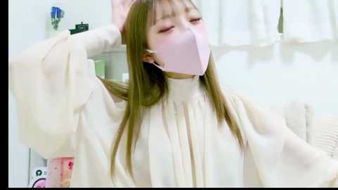 Media: A video of a young woman with long, straight brown hair, wearing a pink mask, white blouse, and a beige headband, stretching her arm. She's in a white room with a pink and white background.