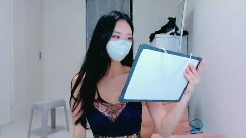 Video of an Asian woman with long black hair, wearing a blue face mask and a black lace bra, holding a clipboard, in a plain, white-walled room with a chair and laundry basket.
