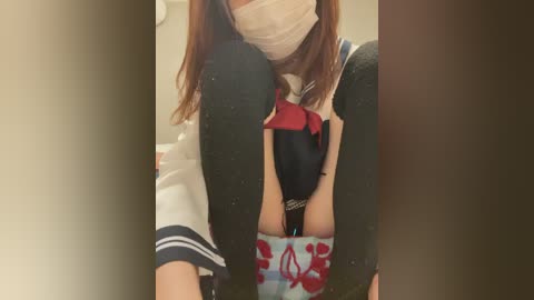 A video of a woman wearing a surgical mask, black leggings, and a white sailor top with a red bow. She is sitting on a blue cushion with floral patterns.